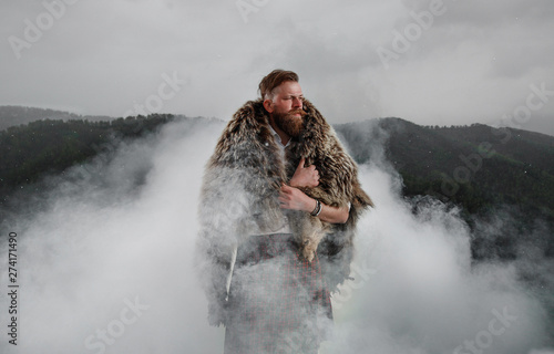 a man with a beard mustache is standing on top of a kilt mountain. bear skin on the back. in a white shirt, with a tattoo on his arm. on the grass. it's snowing. smoke is around