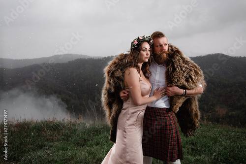 man with beard mustache is standing on top kilt mountain. bear skin back. white shirt, with tattoo on his arm. it is snowing around. smoke. hugs girl wreath cream dress
