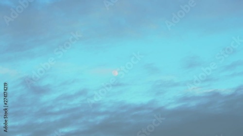 Zooming out on a small moon in the sky surrounded by clouds, taken early evening