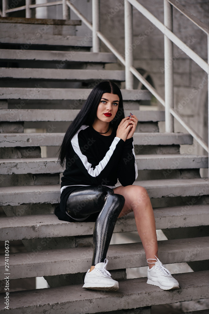 Portrait of attractive handicapped woman in black tracksuit with prosthetic  leg sitting at the street stairs and holding thermos cup Stock Photo