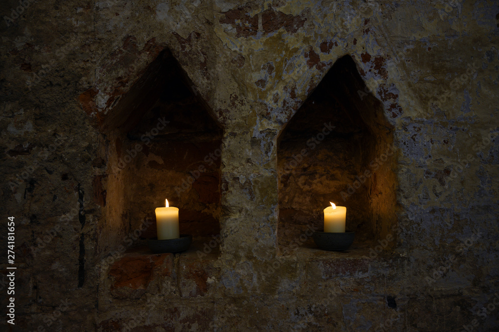 Fototapeta premium two white burning candles in two niches in a dark old brick wall of a monastery, copy space