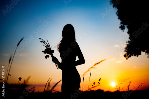 Silhouette of slim girl with a bouquet of flowers. girl stands at sunset in the field