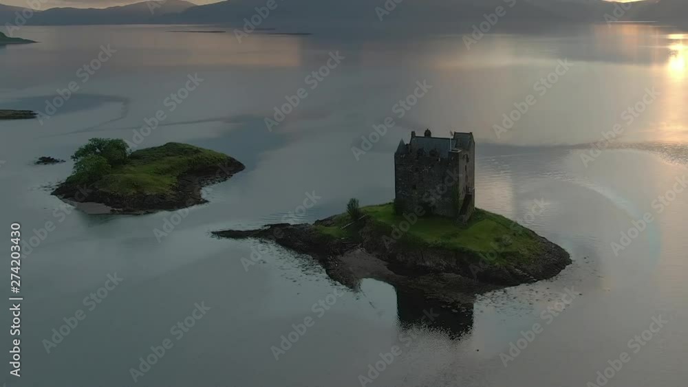 An aerial view of Castle Stalker on Loch Laich as the sun begins to set. Flying right to left and zooming out, whilst descending with mountains and the setting sun behind the castle. An aerial view of Castle Stalker on Loch Laich as the sun begins to set. Flying right to left and zooming out, whilst descending with mountains and the setting sun behind the castle.