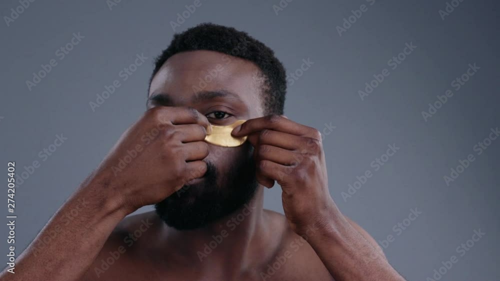 Beautiful sensual young black man applying golden collagen mask on facial skin. Portrait of african topless sexy guy preparing for the day using eye patches looking straight on grey background.