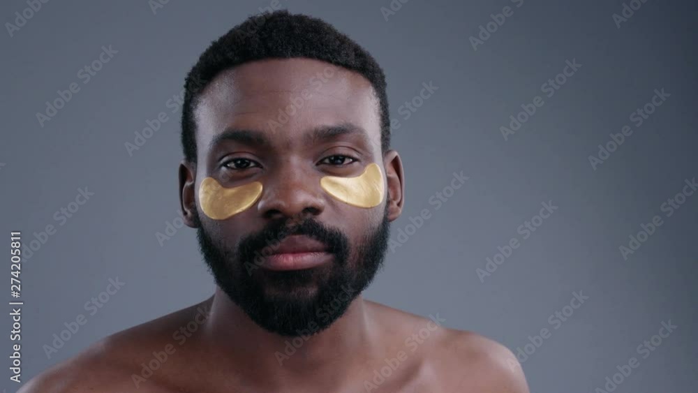 Portrait of mindful beauty afro-american man with under eye patches isolated on grey background. Close-up of sexy male model having spa facial treatment looking at camera. Portrait of mindful beauty afro-american man with under eye patches isolated on grey background. Close-up of sexy male model having spa facial treatment looking at camera.