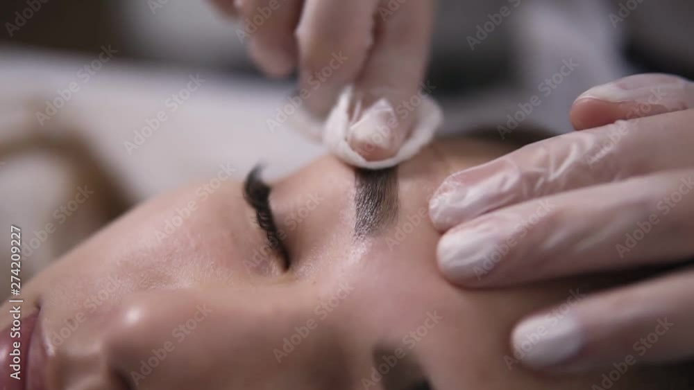 Using a white clean cotton disk to remove the paint from the brows. A woman lay on the bed during a visit at the beauty salon Using a white clean cotton disk to remove the paint from the brows. A woman lay on the bed during a visit at the beauty salon