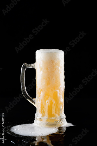overflowing mug of fresh cold light beer with froth flowing on the table and beautiful spilled beer on a black background with reflection