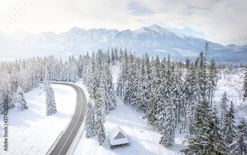 Snowy winter mountains landscape with frosty forest and road. Road to rocky mountain range. Wooden house in pine trees in mountain village