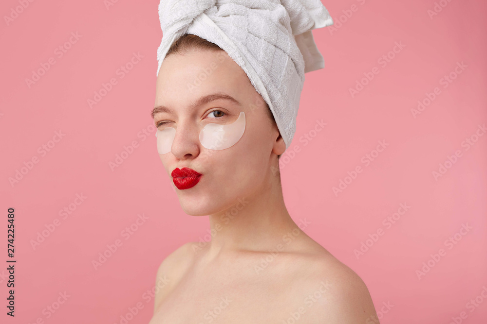 Close up of young woman with a towel on her head after shower, with patches and red lips, looks happy, sends kiss and stands over pink background. Close up of young woman with a towel on her head after shower, with patches and red lips, looks happy, sends kiss and stands over pink background.