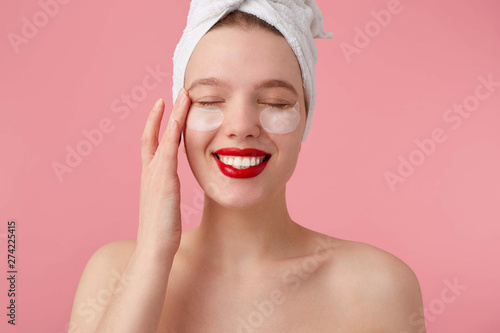 Portrait of young enjoying woman after shower with a towel on her head, with patches and red lips, touches face, stands over pink background.