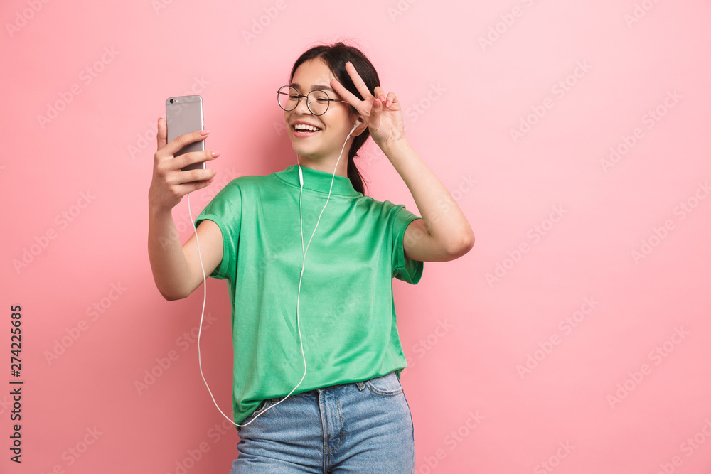 Photo of joyful young girl wearing round eyeglasses taking selfie on mobile phone while using earphones Photo of joyful young girl wearing round eyeglasses taking selfie on mobile phone while using earphones