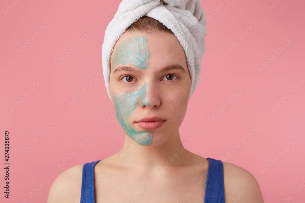 Close up of young nice lady with a towel on her head after shower, put a mask on the floor to compare the effect, looks at the camera stands over pink background Close up of young nice lady with a towel on her head after shower, put a mask on the floor to compare the effect, looks at the camera stands over pink background