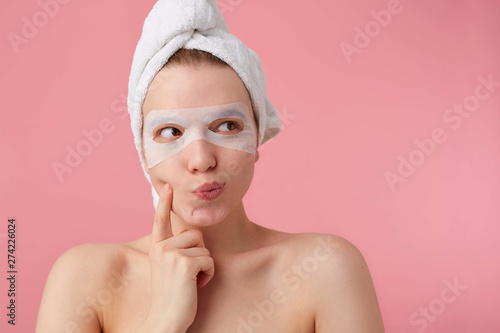 Close up of thinking young woman with a towel on her head after shower, looks away and touches cheek, can't make a decision, stands over pink background.