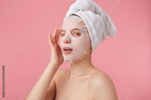 Portrait of young positive smiling woman after spa with a towel on her head, with mask for face, enjoys for time for self care, winks, looks at the camera stands over pink background.