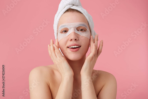Close up of young smiling happy woman after spa with a towel on her head, with mask for eyes, broadly smiles, looks positive, stands over pink background.