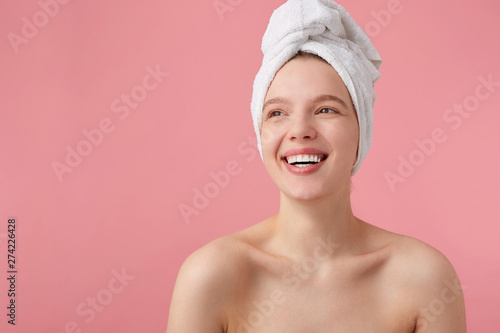 Studio photo of cheerful young lady after shower with a towel on her head, broadly smiles, looks away, feels so happy, stands over pink background.