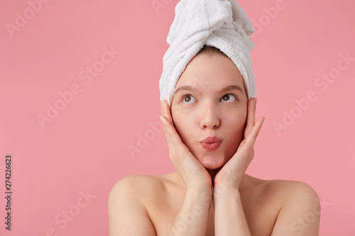 Close up of young happy woman after shower with a towel on her head, dreamily looking away with palms on cheeks, standing over pink background.
