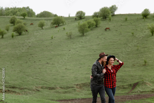 Fototapeta Naklejka Na Ścianę i Meble -  a man with a beard and mustache in a leather jacket and a woman in a plaid shirt in hats are holding hands, on green hills, a ranch. horses are walking.caucasian
