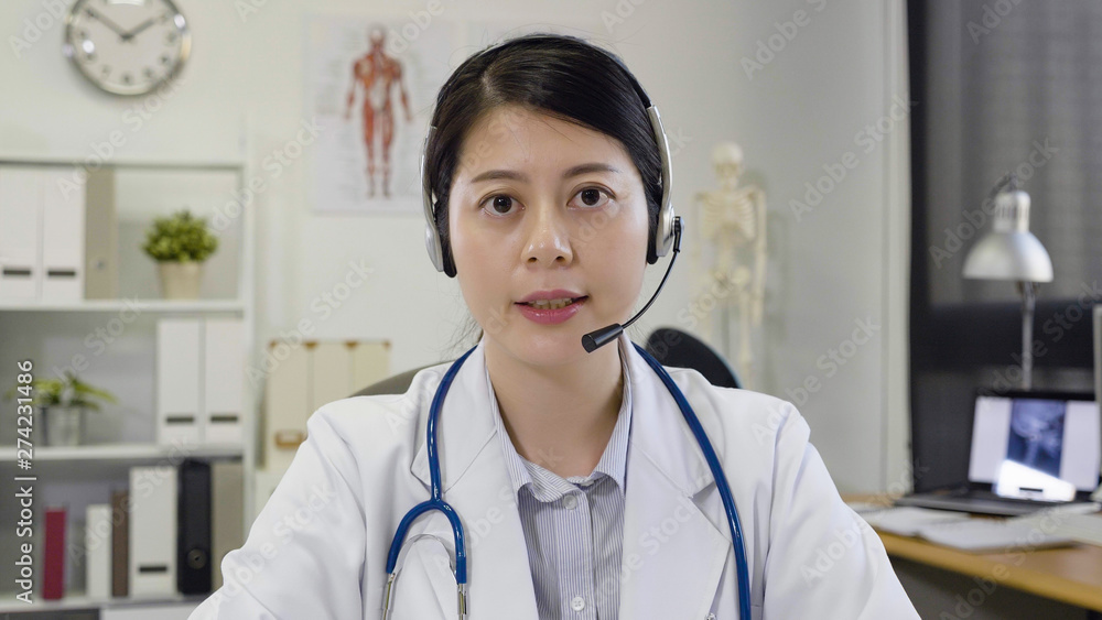 Young asian woman doctor with stethoscope and headphones in white uniform sitting at working desk in clinic office face camera talking on service center with patient. medical nurse answer phone call Young asian woman doctor with stethoscope and headphones in white uniform sitting at working desk in clinic office face camera talking on service center with patient. medical nurse answer phone call