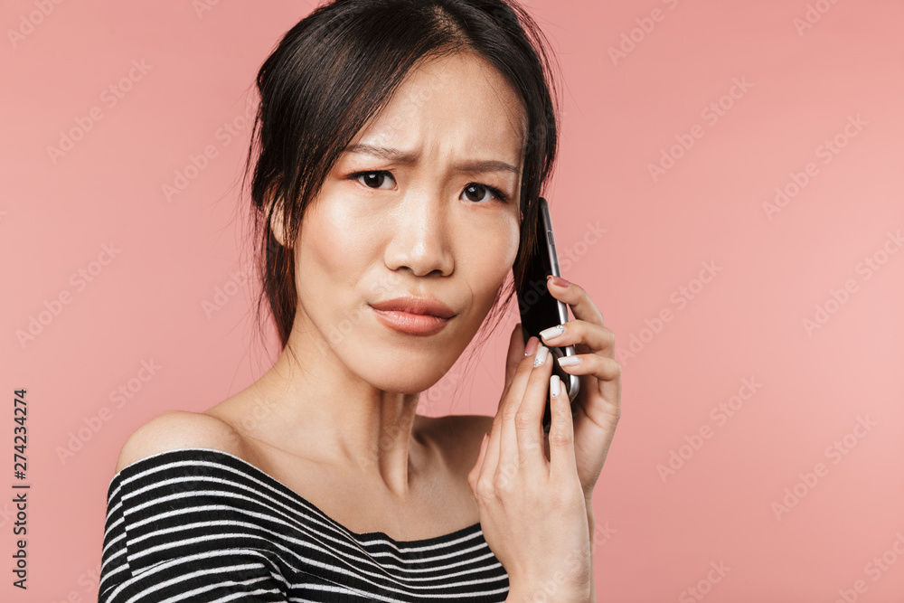 Photo of disappointed asian woman dressed in basic wear frowning and talking on smartphone Photo of disappointed asian woman dressed in basic wear frowning and talking on smartphone