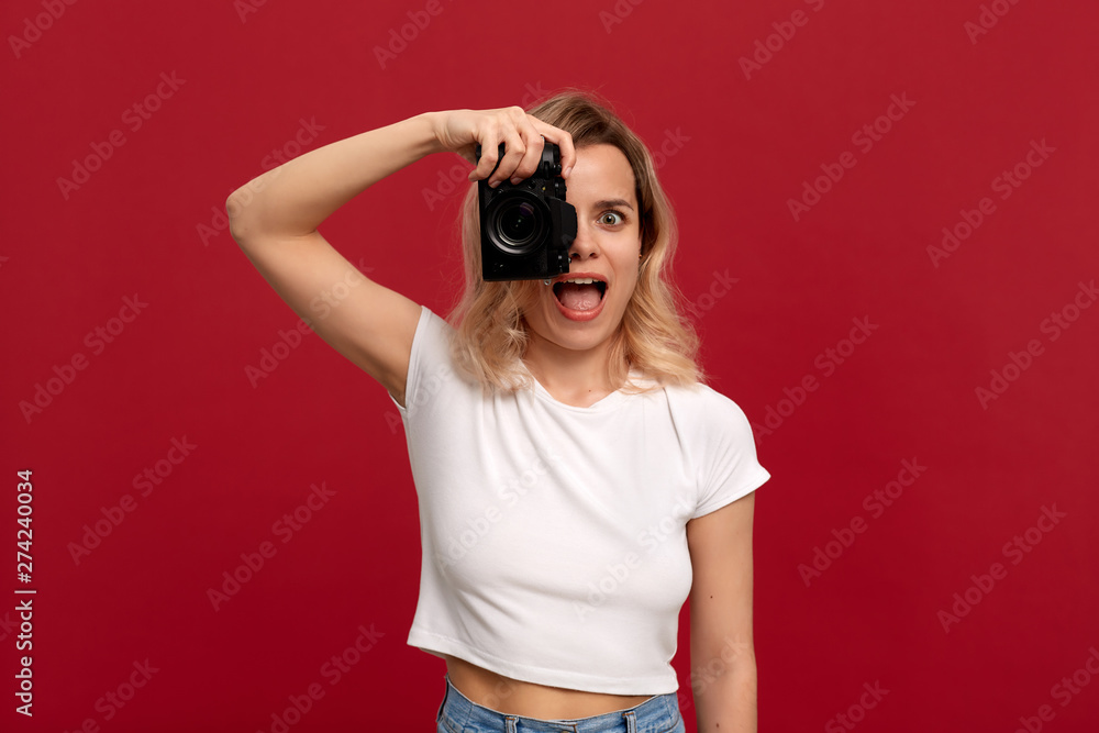 Portrait of a girl with curly blond hair dressed in a white t-shirt standing on a red background. Model takes photos with retro camera. Portrait of a girl with curly blond hair dressed in a white t-shirt standing on a red background. Model takes photos with retro camera.