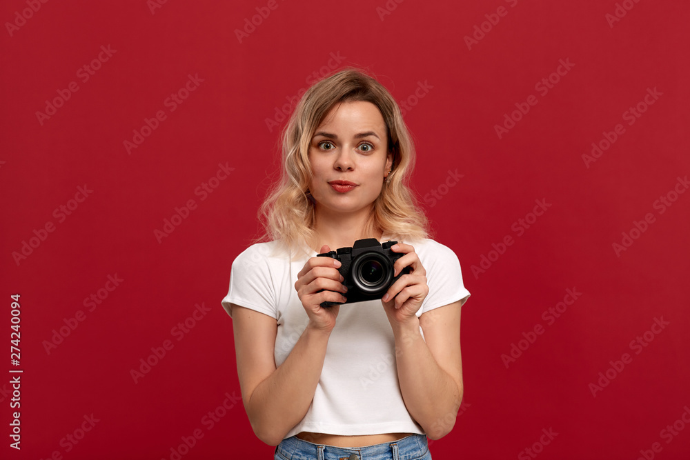 Portrait of a girl with curly blond hair dressed in a white t-shirt standing on a red background. Model takes photos with retro camera. Portrait of a girl with curly blond hair dressed in a white t-shirt standing on a red background. Model takes photos with retro camera.
