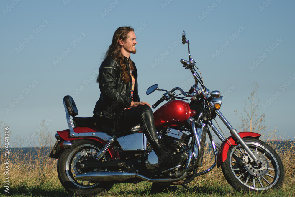 Fototapeta premium Przystojny rowerzysta długowłosy w skórze na czerwonym motocyklu z pola i brzegu morza. Brutalny mężczyzna w skórze