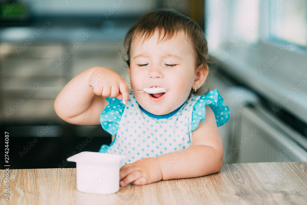 Cute, funny girl eating yogurt in the kitchen in a blue dress or apron in the afternoon Cute, funny girl eating yogurt in the kitchen in a blue dress or apron in the afternoon