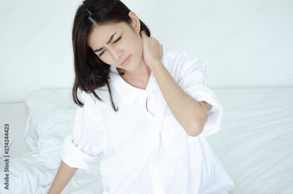 Asian women wear white pajamas in the bedroom. She has pain in the neck area.Do not focus on the main object of this image.