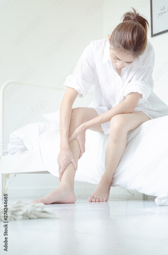 Asian girl wearing white dress have leg pain. The woman was squeezing her leg in a white bedroom with sunshine.Warm tone.Do not focus on objects. Asian girl wearing white dress have leg pain. The woman was squeezing her leg in a white bedroom with sunshine.Warm tone.Do not focus on objects.