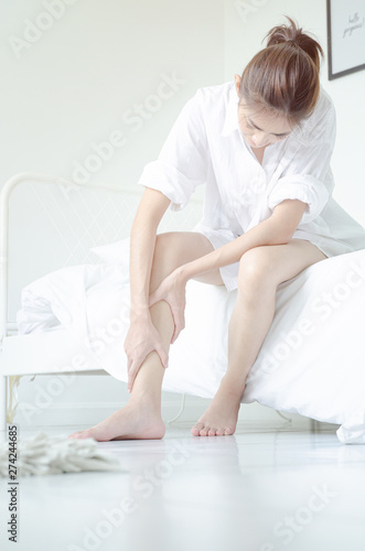 Asian girl wearing white dress have leg pain. The woman was squeezing her leg in a white bedroom with sunshine.Warm tone.Do not focus on objects.