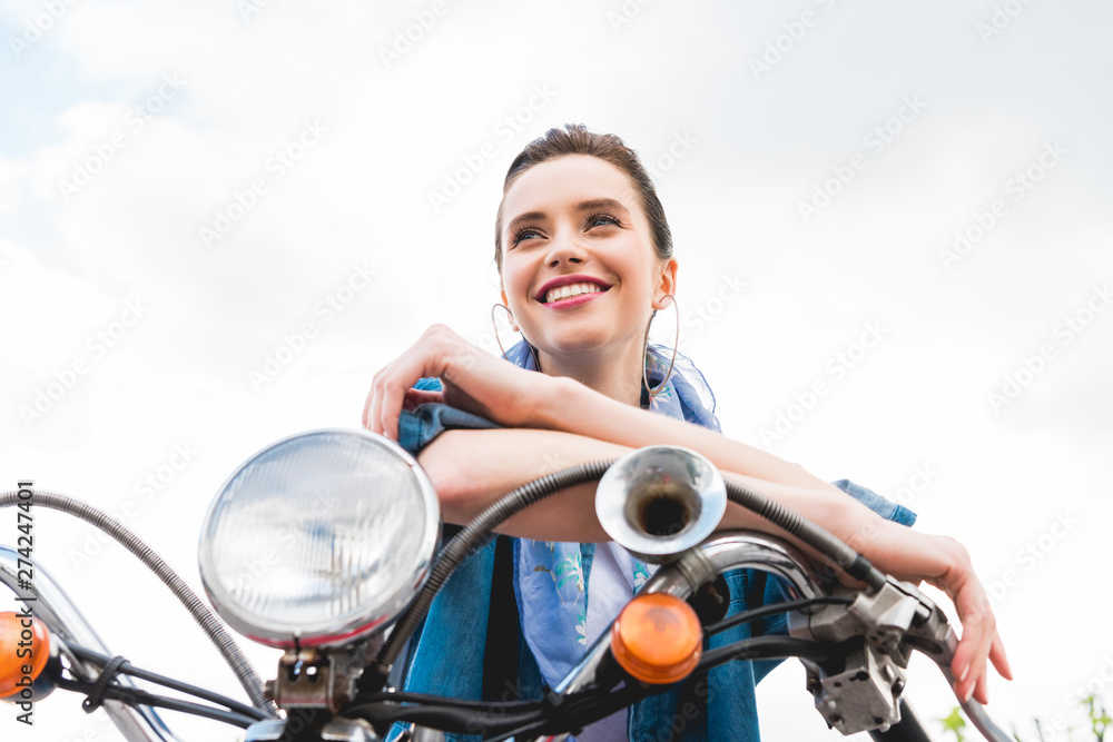 low angle view of beautiful girl sitting on scooter, resting, looking away and smiling on sky background