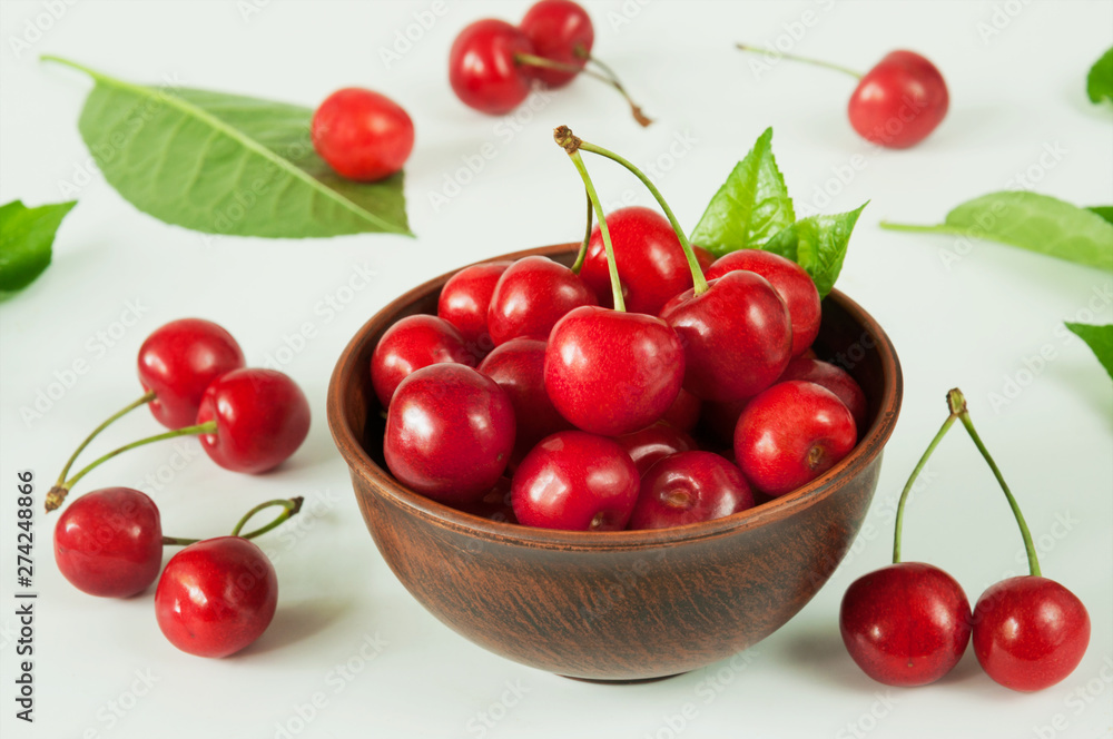 Close-up of ripe red sweet cherry with branches and leaves in a ceramic brown bowl on a white background. Selective focus. Close-up of ripe red sweet cherry with branches and leaves in a ceramic brown bowl on a white background. Selective focus.
