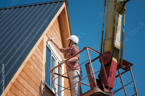 a young woman paints the window frames of a wooden house on the top floor with a aerial platform