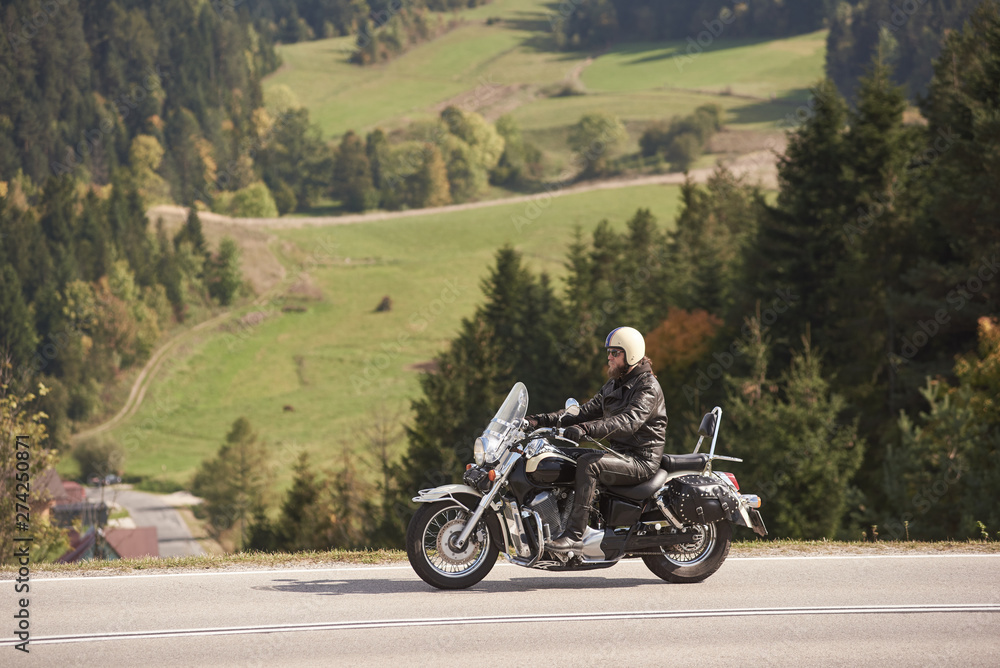 Side view of bearded motorcyclist in helmet, sunglasses and black leather clothing riding cruiser motorbike along narrow asphalt path on sunny summer day on background of tall trees and greeen grass.