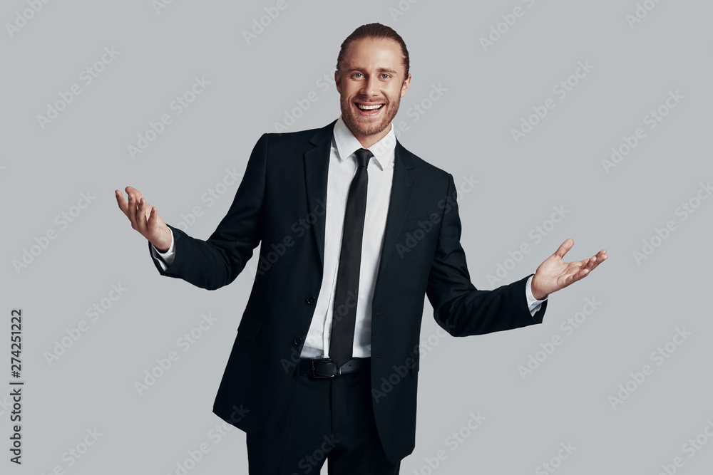 Perfect in every way. Handsome young man in full suit looking at camera and keeping arms outstretched while standing against grey background Perfect in every way. Handsome young man in full suit looking at camera and keeping arms outstretched while standing against grey background