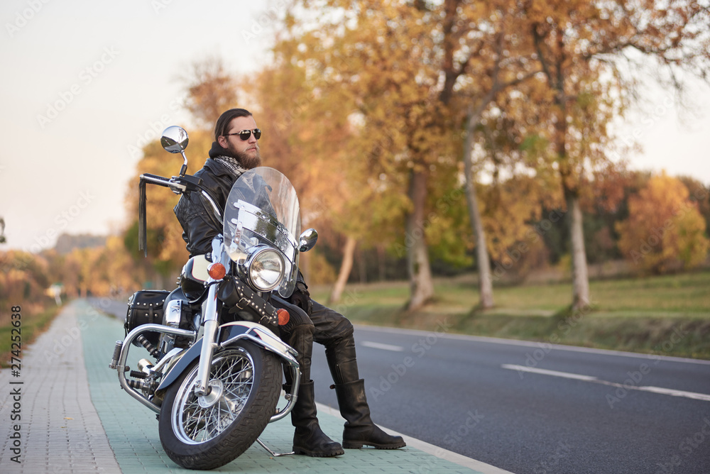 Young bearded biker in black leather clothing and dark sunglasses sitting on motorcycle on clean paved roadside, on background of empty straight asphalt road and vintage trees golden bokeh foliage.