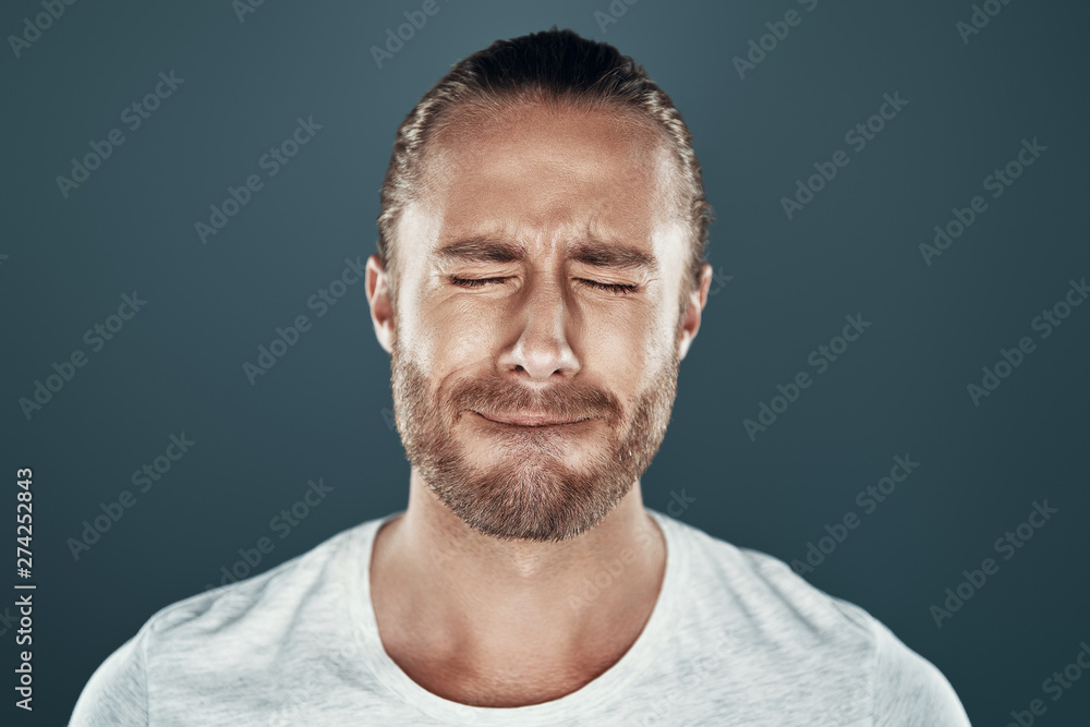 Fototapeta premium Hate to lose. Sad young man keeping eyes closed and making a face while standing against grey background