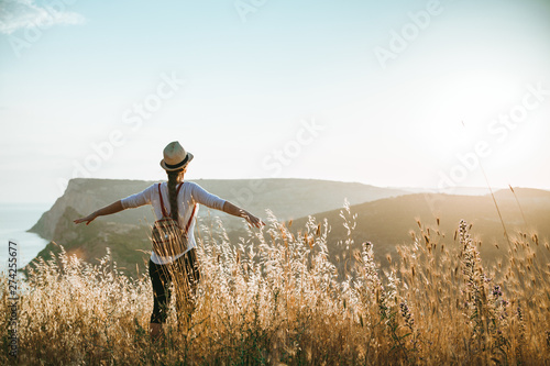 A young girl stands with a hat and a backpack against the backdrop of mountains bathed in the rays of the sun. Summer background with place for your text