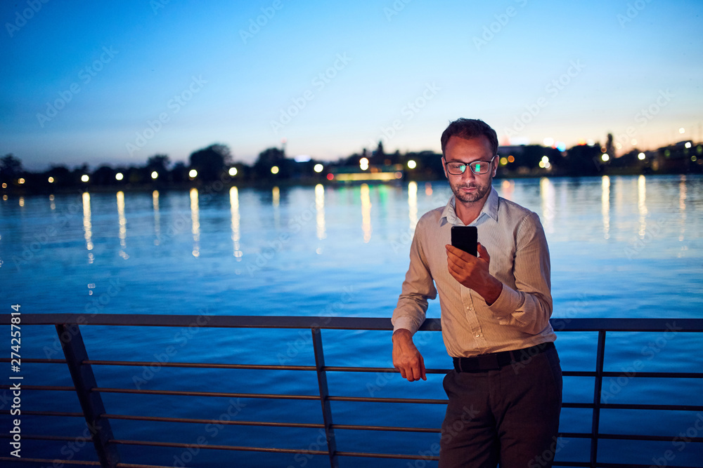 Businessman using cellphone near the river with city lights in background.