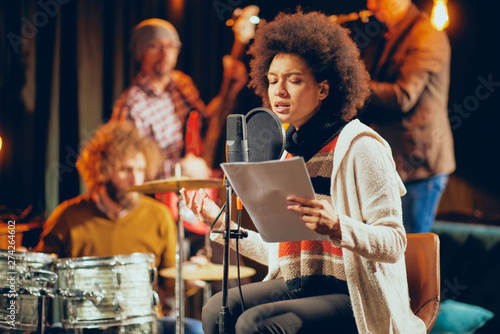 Mixed race woman singing. In background band playing instruments. Home studio interior.