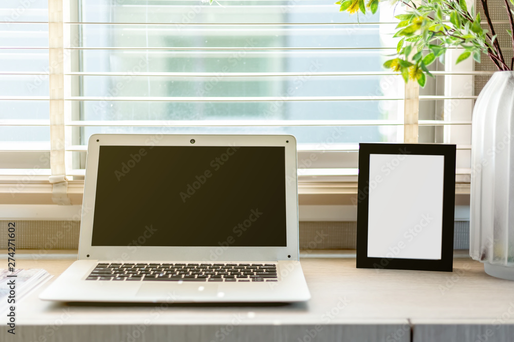 Fototapeta premium Blank black color picture frame template for place image or text inside on the desktop table work space with laptop and flower. window light