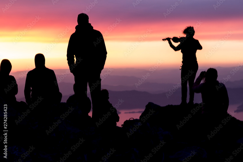 Multi-colored sunset. Silhouette of a woman with a violin. Silhouettes of people on top of a mountain overlooking the sea.
