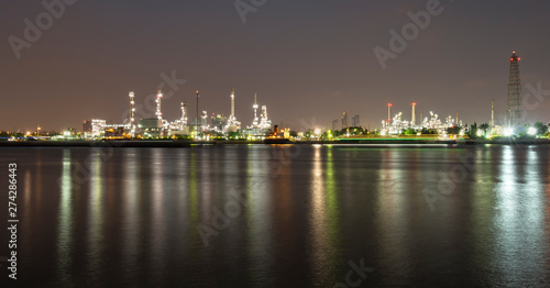 Long exposure night photo crude oil refinery plant and many chimney with petrochemical tanker or cargo ship at coast of the river with colorful bright light from lamp reflect on water at thailand