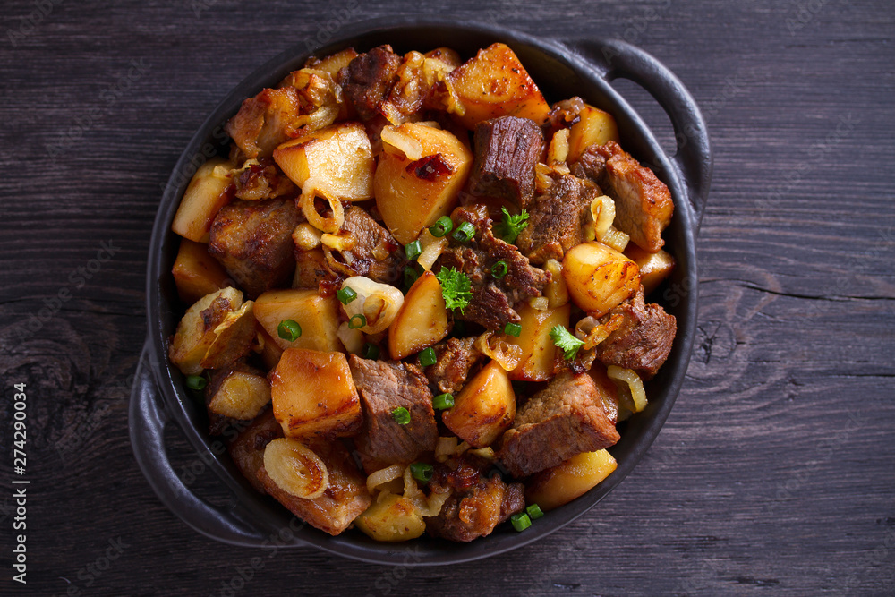 Fried beef and potatoes with onions and garlic served in black dish on wooden background Fried beef and potatoes with onions and garlic served in black dish on wooden background