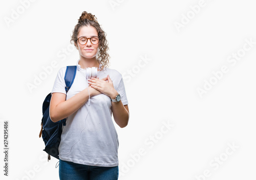 Young brunette student girl wearing backpack and headphones over isolated background smiling with hands on chest with closed eyes and grateful gesture on face. Health concept.