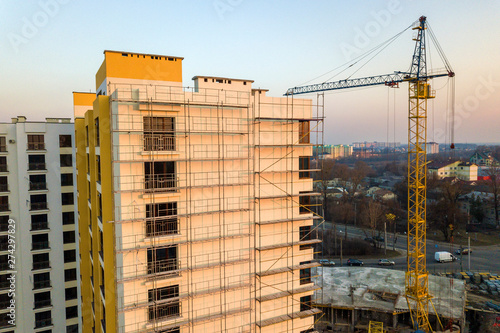 Apartment or office tall unfinished building under construction. Brick wall in scaffolding, shiny windows and tower crane on urban landscape and blue sky background. Drone aerial photography. © bilanol