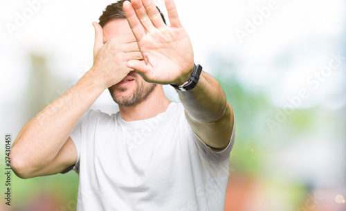 Handsome man wearing casual white t-shirt covering eyes with hands and doing stop gesture with sad and fear expression. Embarrassed and negative concept.