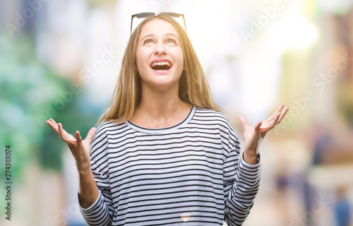 Young beautiful blonde woman wearing sunglasses over isolated background crazy and mad shouting and yelling with aggressive expression and arms raised. Frustration concept.