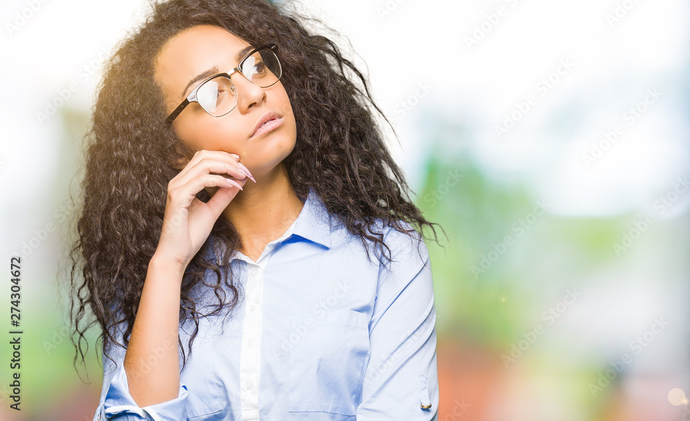 Young beautiful business girl with curly hair wearing glasses with hand on chin thinking about question, pensive expression. Smiling with thoughtful face. Doubt concept. Young beautiful business girl with curly hair wearing glasses with hand on chin thinking about question, pensive expression. Smiling with thoughtful face. Doubt concept.